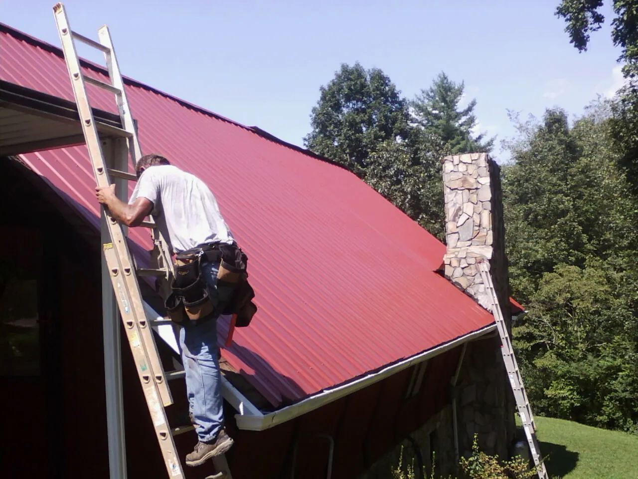 Roofer on a ladder beside a red metal roof and stone chimney — Soffit & Fascia Repair by Roofing Brothers in Sonyea